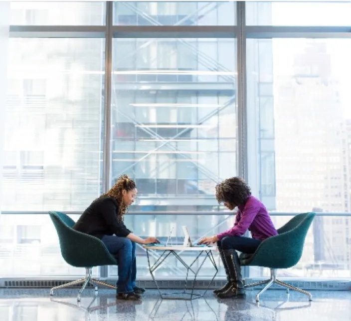 Two women talking in green swivel lounge chairs set across a small round table in front of floor-to-ceiling windows.