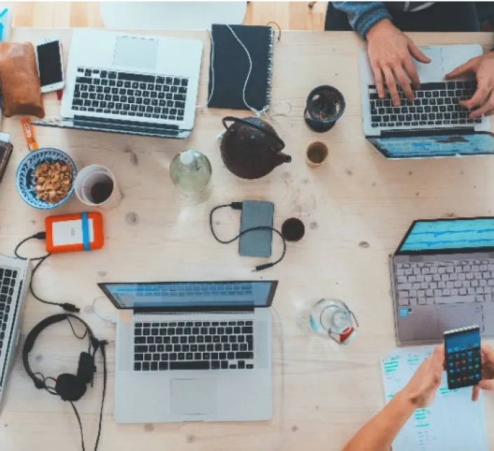 Overhead view of a light wooden table shared by several people working on laptops, with a teapot, headphones and phones between them.